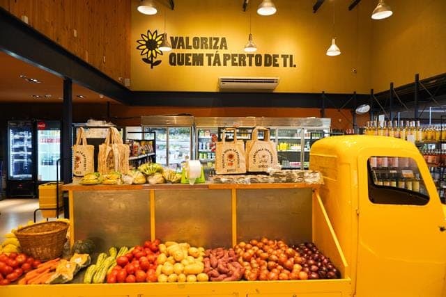 Hortifruti do Brasco Viva montado num tuk-tuk amarelo, sob a parede com a frase Valoriza quem tá perto de ti.